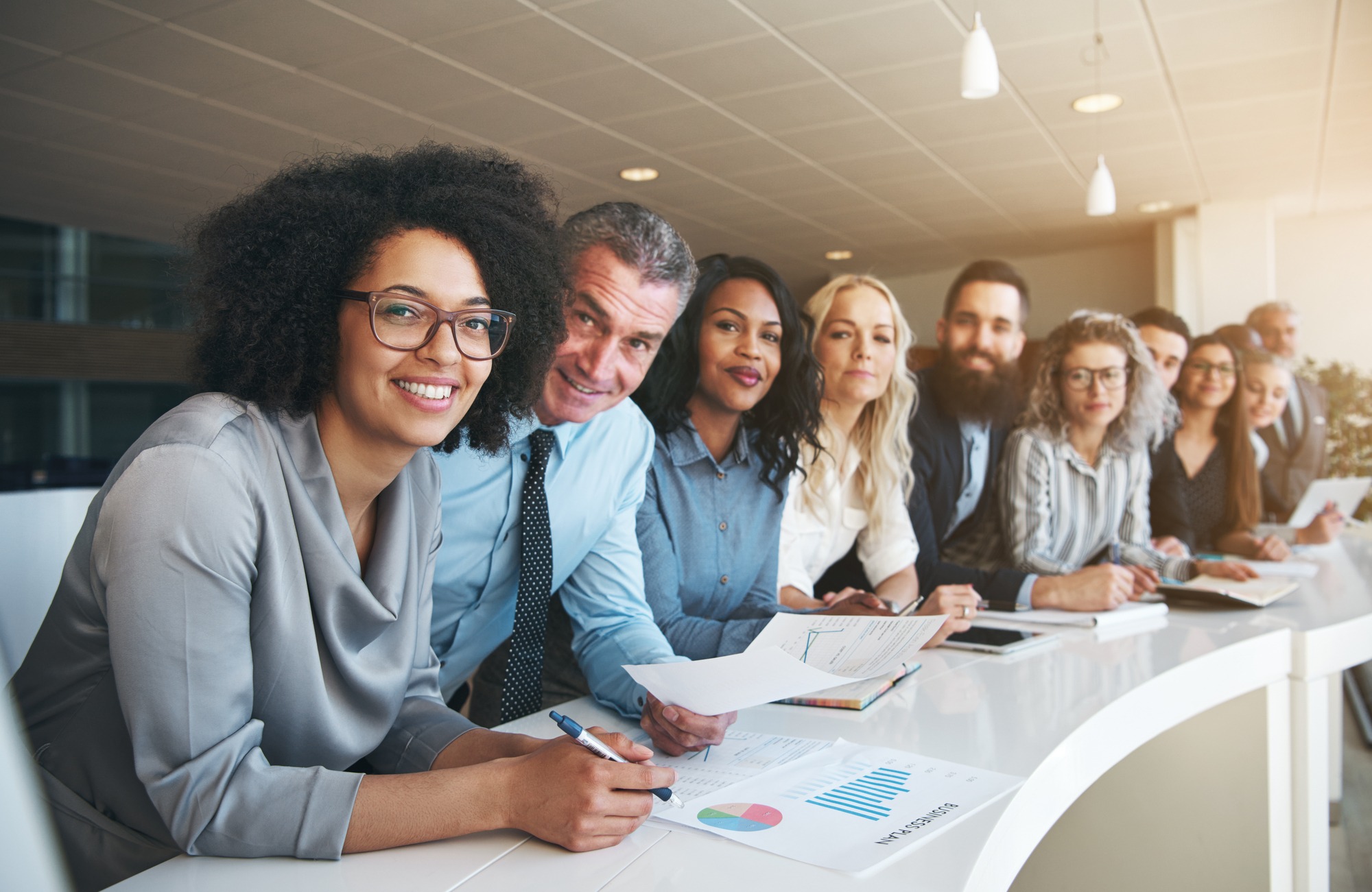 Portrait of a smiling group of diverse corporate colleagues standing in a row together at a table in a bright modern office.